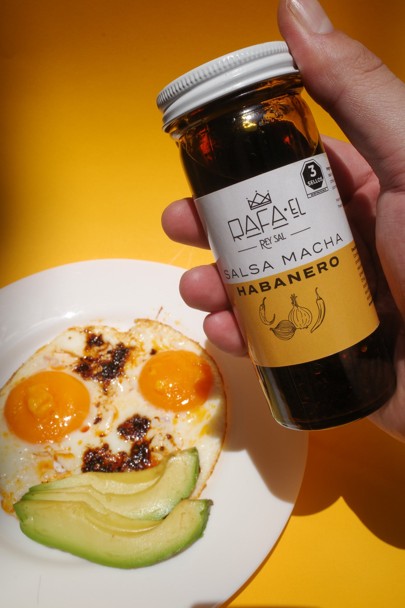 A person holding a bottle of salsa. The bottle has a white lid with black and white numbers on it. On the table next to them is a plate of food consisting of two eggs with salsa macha and an avocado. The light in the room casts shadows across the dish. This scene conveys warmth and comfort from being surrounded by familiar objects while preparing something delicious to eat.